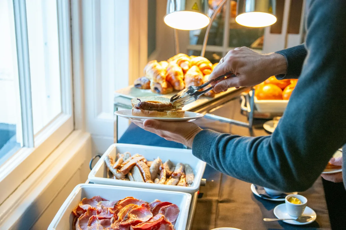 A person uses tongs to take a toasted roll from a breakfast spread of croissants, sausages, bacon and oranges next to a window.