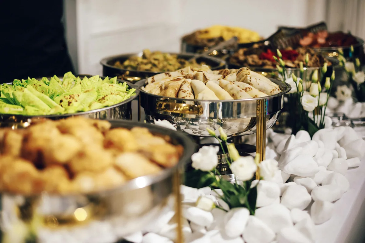 Polished chafing dishes of filled crepes, salad and baked dishes on a white buffet table decorated with white flowers.