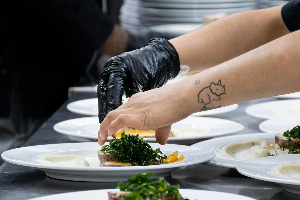 A cook in a black glove tops a long row of identical white plates with kale salad and sliced roast meat in a restaurant kitchen.