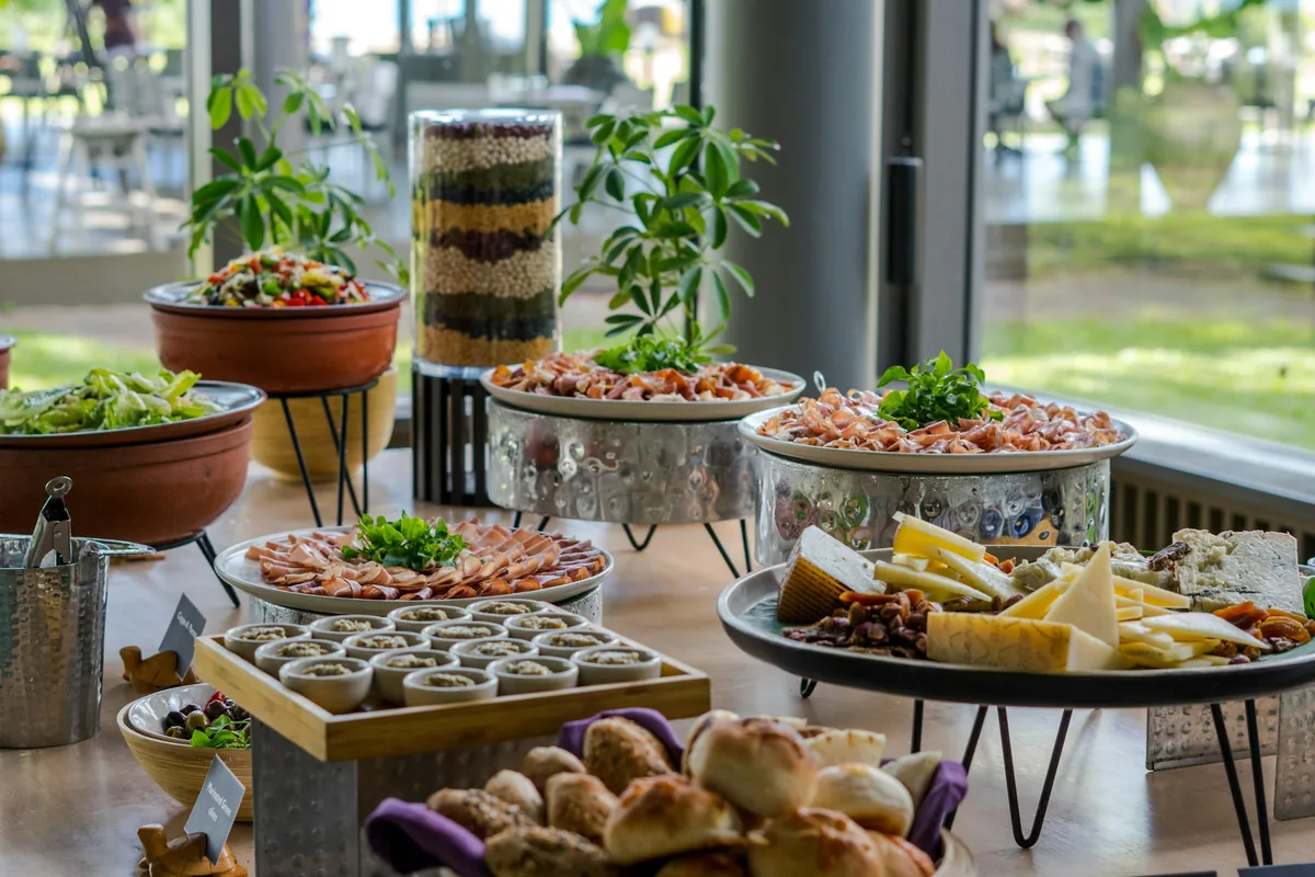 A party spread by a sunny window: a cheese board, platters of cured meats, small bowls of dip, a basket of bread rolls and salads on raised stands.